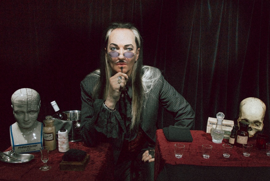 Magician holding glasses to his face, surrounded by alchemical paraphernalia incuding skull, glass vials and bottles.