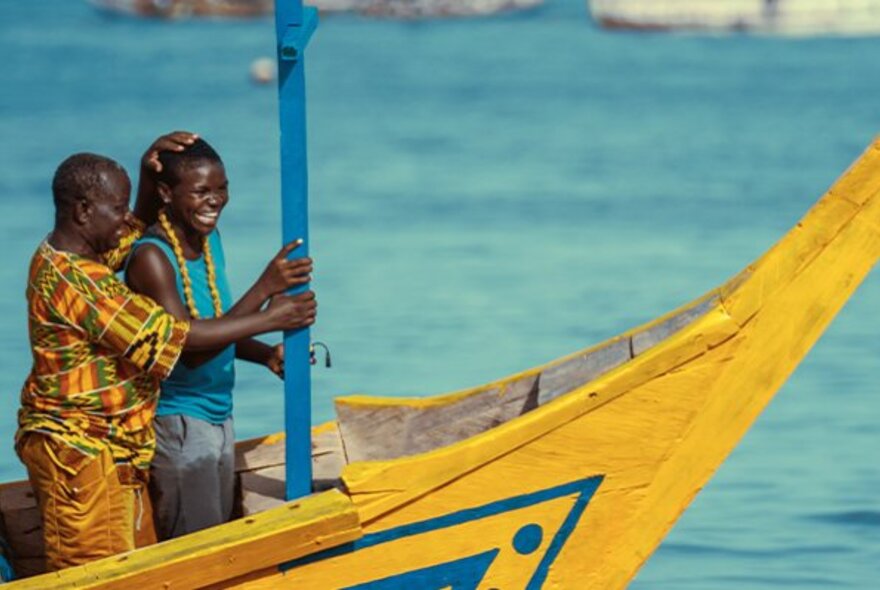 A still from an African film showing two colourfully dressed people standing in the bow of a yellow-painted wooden boat; blue ocean behind them.