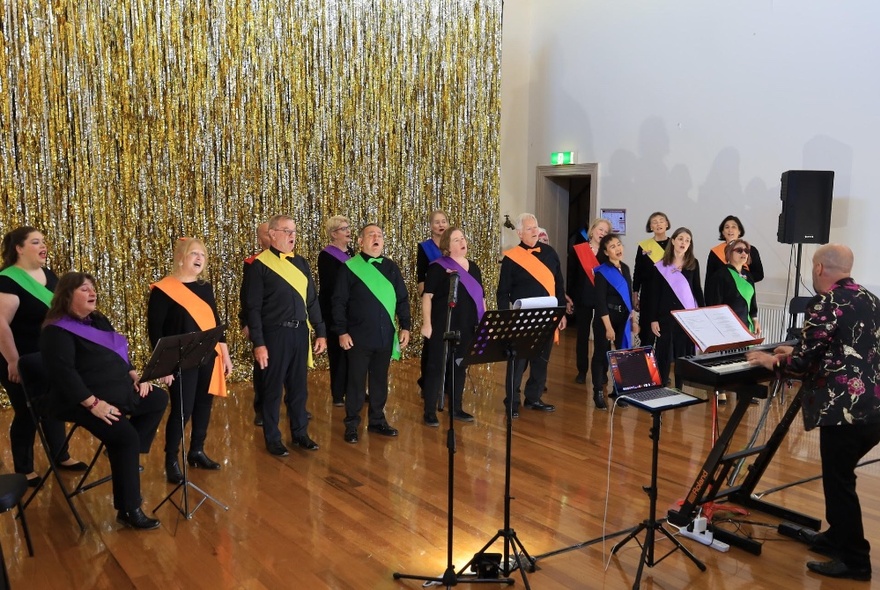 A conductor with a choir performing in a small space, each wearing black with a different coloured sash.