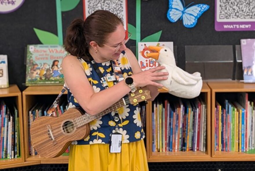 A woman in a library setting holding a ukulele and talking to a toy bird. 