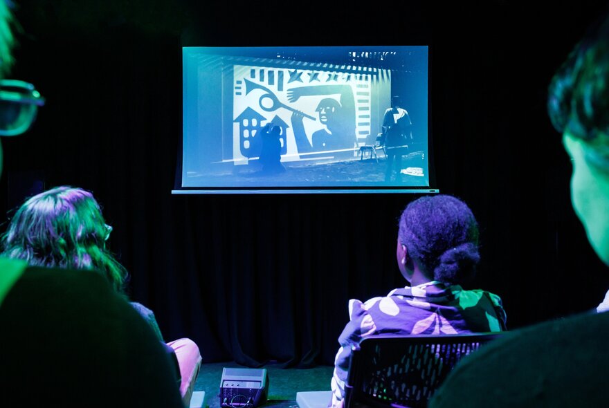 A seated audience in a darkened cinema watching a small cinema screen.