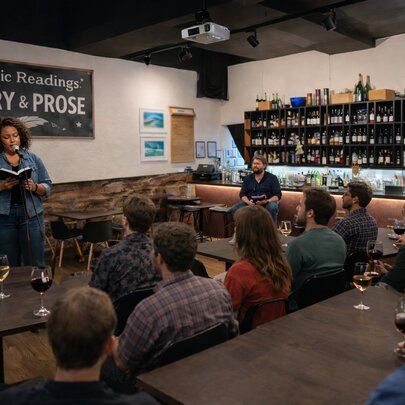 A small gathering in a wine bar with a woman reading under a 'poetry and prose' sign. 
