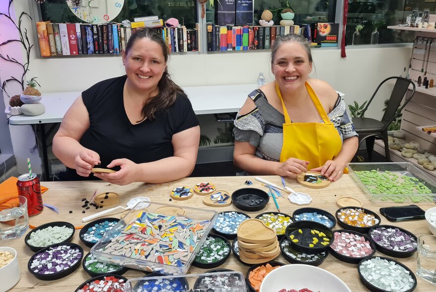 Two workshop participants smiling and seated at a workbench making multicoloured mosaic tiled coasters.