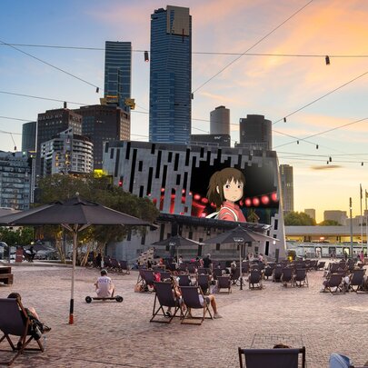 Federation Square Melbourne at dusk with deck chairs out and a Studio Ghibli film showing on the big screen. 