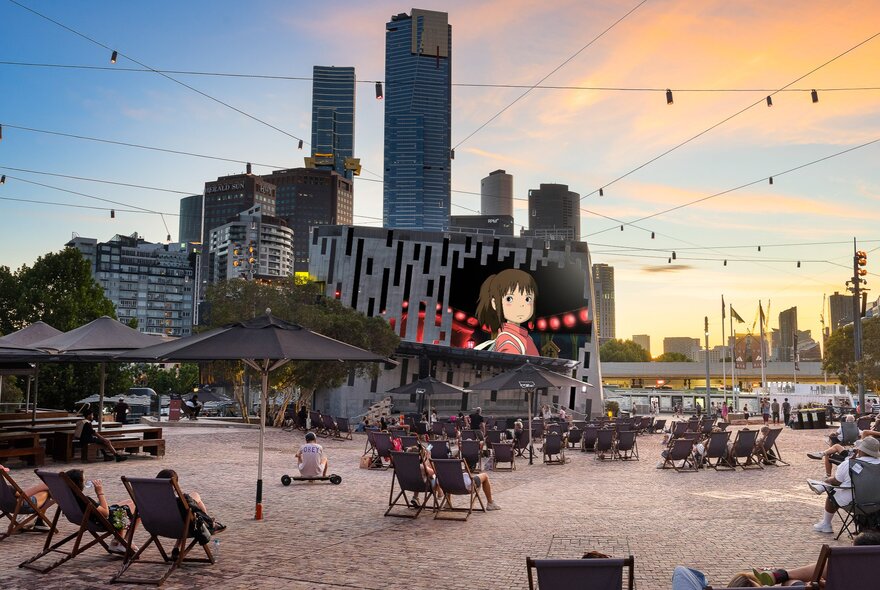 Federation Square Melbourne at dusk with deck chairs out and a Studio Ghibli film showing on the big screen. 
