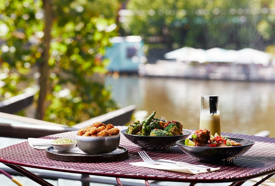 An outdoor restaurant table beside the river, set with three dishes and a cocktail.