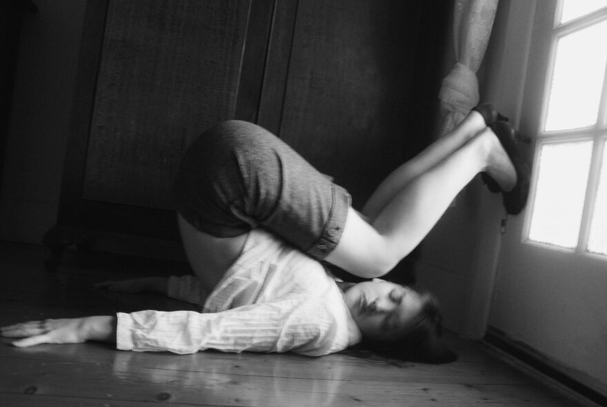 Woman lying on her back, her legs bent over her head, feet against a window.