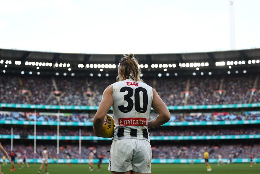 Back view of a Collingwood AFL football player with the ball during a match, with a filled stadium of spectators in the background.