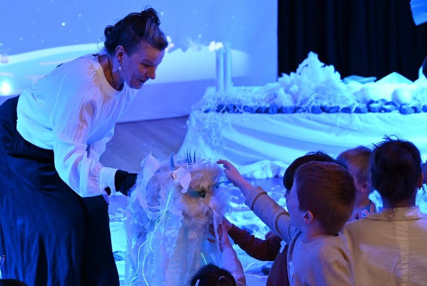 Children lining up to touch something in plastic that a woman is holding out, in an icy blue tinted room. 