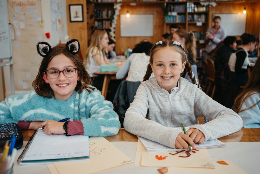 Two young students smiling at the viewer, seated at a table in front of their notebooks, as part of a creative writing workshop in a studio setting.