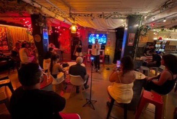 The interior of a bar, with a small patronage seated around a stage where locals are performing stand-up comedy.