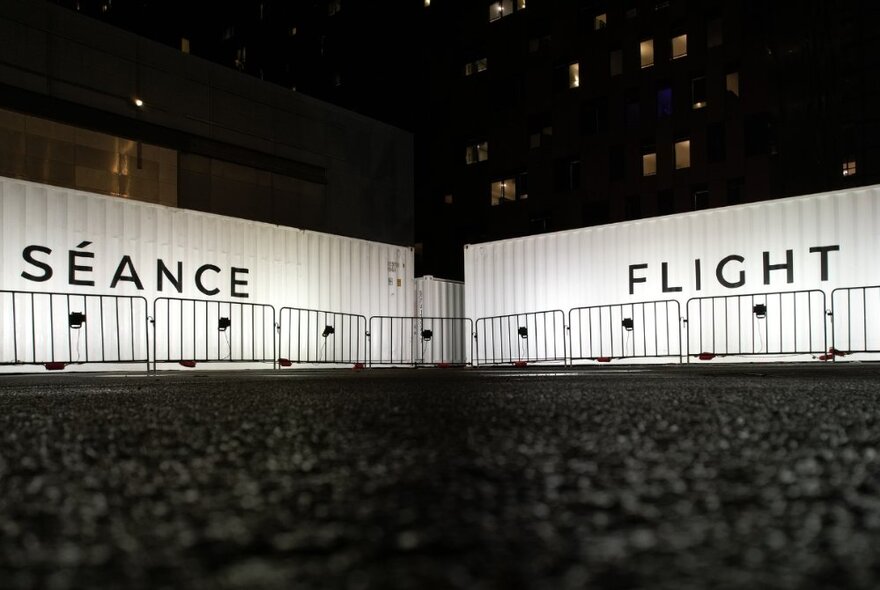 Two white shipping containers in a large open space at night, with the words SEANCE written on one container and FLIGHT written on the other one.