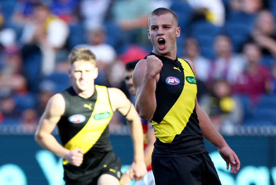 Two Richmond AFL football players on the field, with out-of-focus fans in the stands in the background.