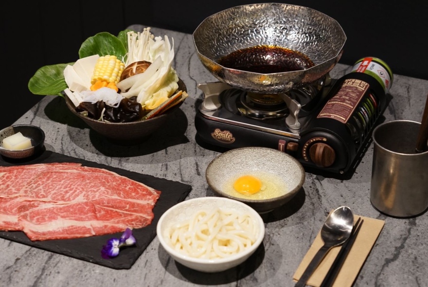A personal Japanese hot pot set up with beef and vegetable ingredients prepared in bowls alongside the small tabletop gas burner with stainless steel bowl of broth.