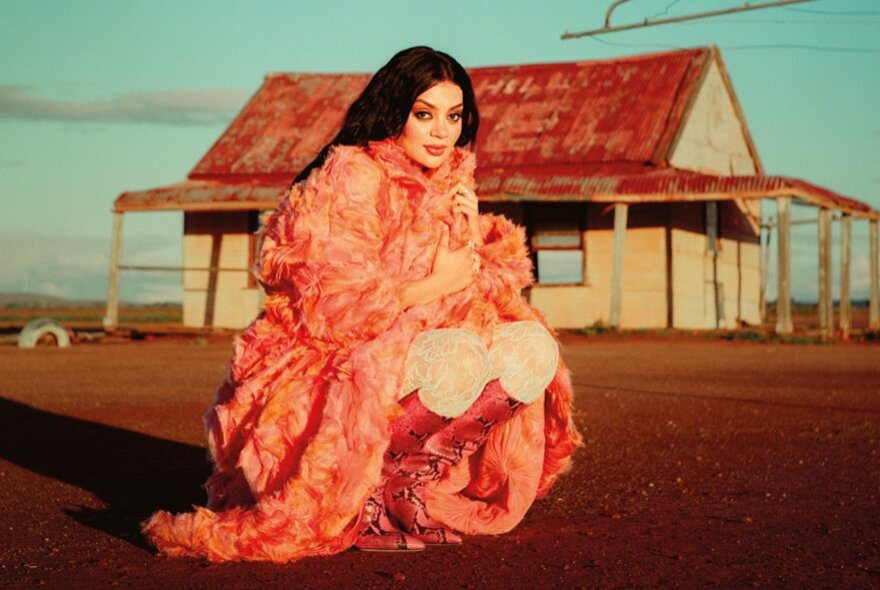 Singer-songwriter Thelma Plum, wearing a voluminous pink dress and boots, squatting on the ground, in an arid desert landscape, with a rustic building in the background.