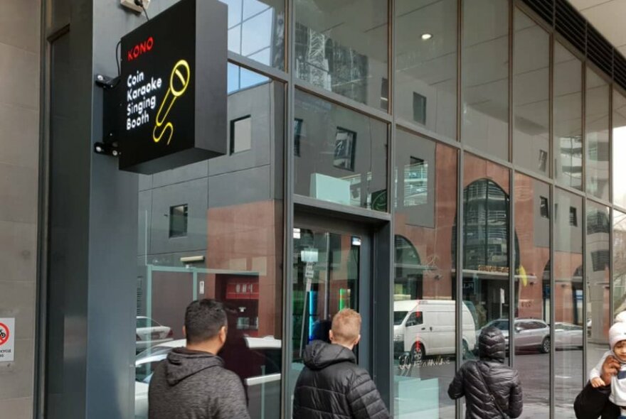 Exterior of building with large glass windows and pedestrians walking past, and signage that reads ' KONO Coin Karaoke Singing Booth'.