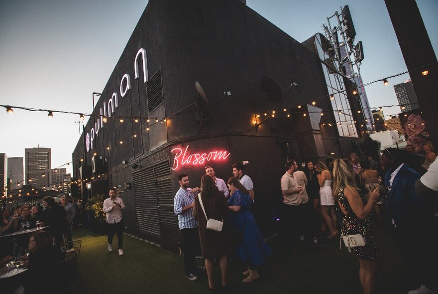 People on a rooftop bar at dusk, fairy lights and a neon Blossom sign.