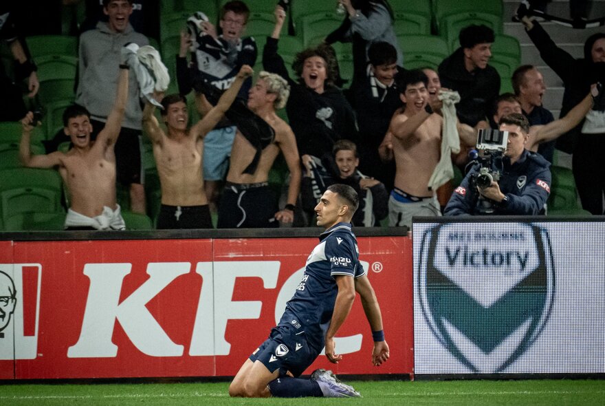 Soccer player on his knees on the grass in front of crowded stands full of male fans baring their chests.