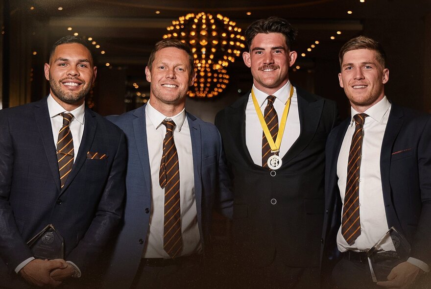 Four smiling men, all wearing suits and matching Hawthorn AFL club ties, posed in a room, one of them with a medal around his neck.