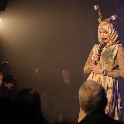 A performer on a stage wearing a costume, talking into a microphone they hold in their hands, in front of a live audience.