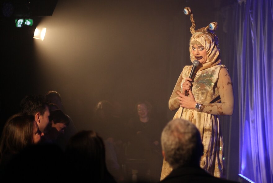 A performer on a stage wearing a costume, talking into a microphone they hold in their hands, in front of a live audience.