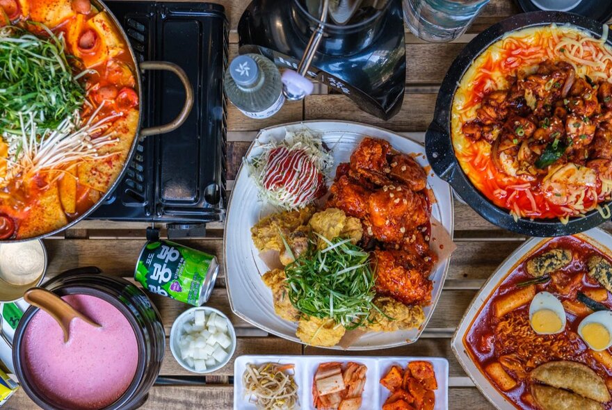 Top down view of plates of food on a wooden table at a Korean restaurant, including fried chicken, curries and noodle dishes.