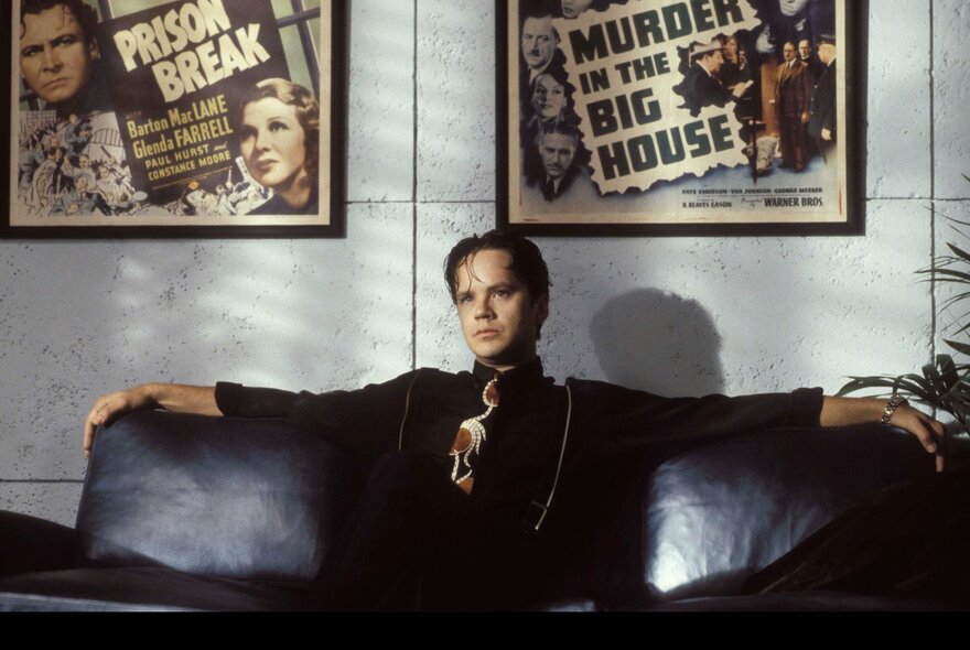 A still from the film, The Player, showing a man in a black shirt and tie, seated on a black sofa with arms outstretched, two framed movie posters hanging on the wall behind him.