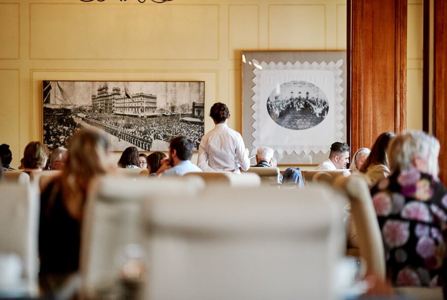 Interior of The Windsor Hotel dining room, with blurred chairs in the foreground and patrons and a waiter in the background.