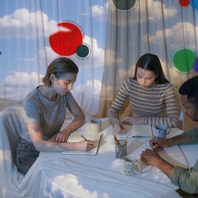 People seated at a table, writing on a table covered with a sky-patterned cloth, sky on curtains in the background.