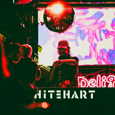 A DJ playing a set inside a darkened Whitehart Bar, featuring a neon 'Delight' sign on the screen behind the DJ and a disco ball overhead.