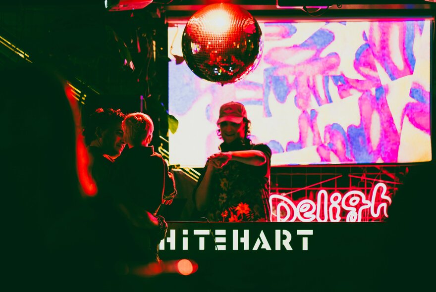 A DJ playing a set inside a darkened Whitehart Bar, featuring a neon 'Delight' sign on the screen behind the DJ and a disco ball overhead.