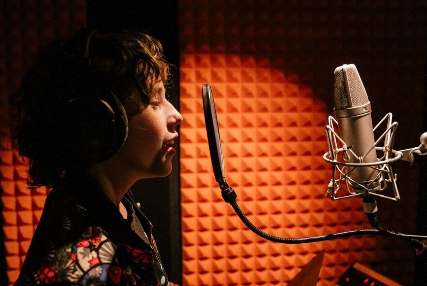 A young teenager in profile wearing headphones and singing into a microphone in a recording studio.