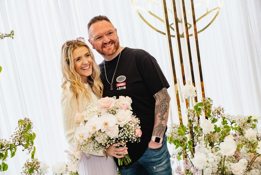 Tattooed man and blonde-haired woman holding a bunch of flowers in a white tent with flowers.