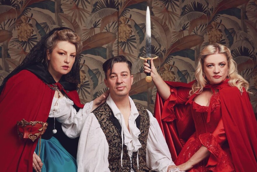 Three cast members of opera production of Don Giovanni, in theatrical costumes posing in front of patterned wallpaper.
