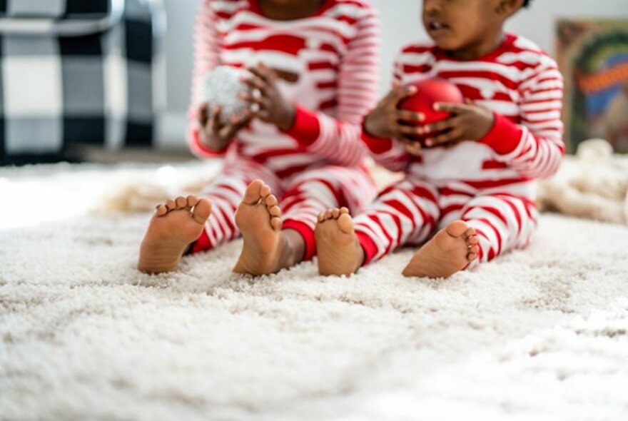 Two young children, both wearing red and white striped onesies, sitting on the floor on a white rug, each holding a Christmas bauble in their hands.