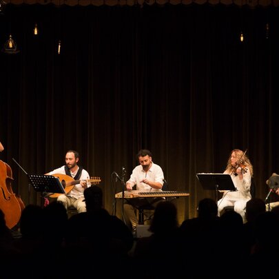 A group of mostly seated musicians perform on a darkened stage. 
