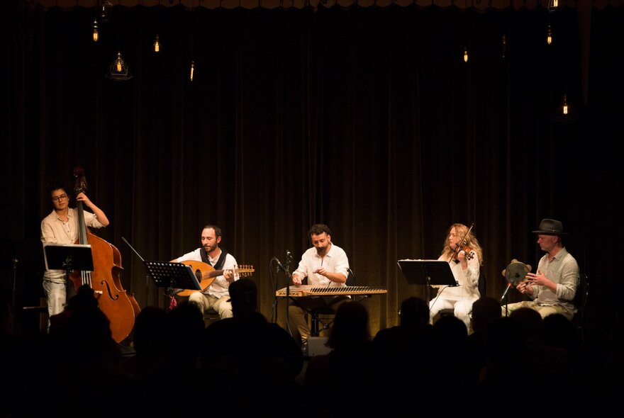 A group of mostly seated musicians perform on a darkened stage. 