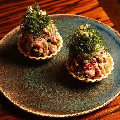 A dish of Akajiso Kingfish presented piled into two scallop shells, with a green seaweed garnish.