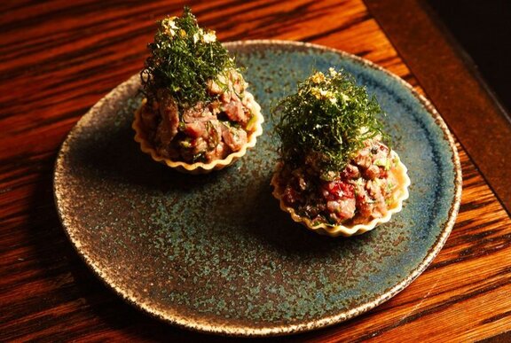 A dish of Akajiso Kingfish presented piled into two scallop shells, with a green seaweed garnish.
