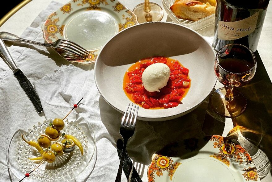 Overhead table setting showing plates of food, a bottle of wine and a wine glass on a white tablecloth.