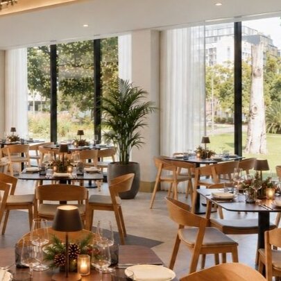 The contemporary restaurant interior of Fico, featuring floor-to-ceiling glass windows that look out onto a lush, green park with mature trees, with tables set up for a dining service.