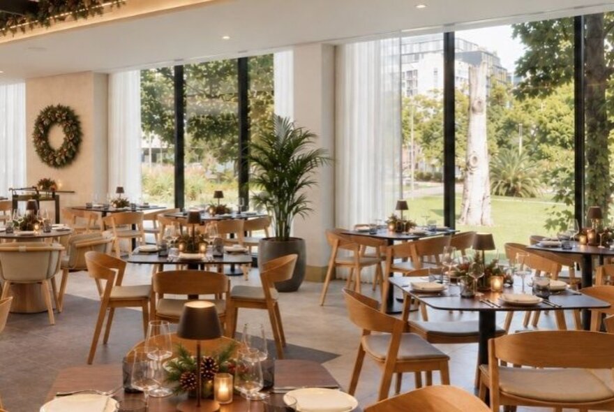 The contemporary restaurant interior of Fico, featuring floor-to-ceiling glass windows that look out onto a lush, green park with mature trees, with tables set up for a dining service.