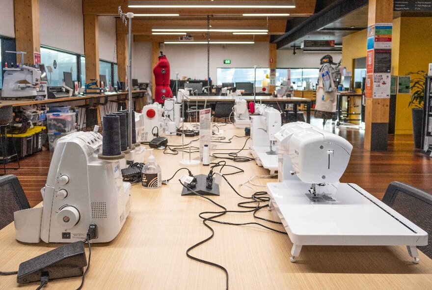 A long table in a large spacious room that has several sewing machines, overlockers and other machines for crafting arranged on it in front of chairs.