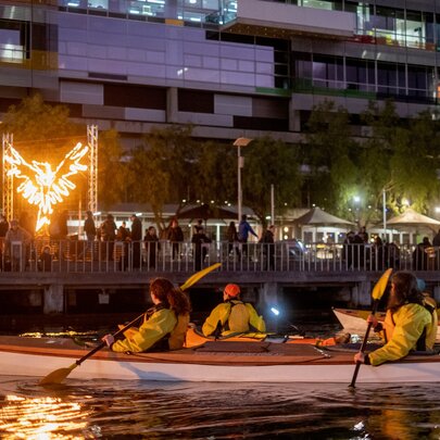People in kayaks on the water next to a busy walkway with a fire installation in the background.