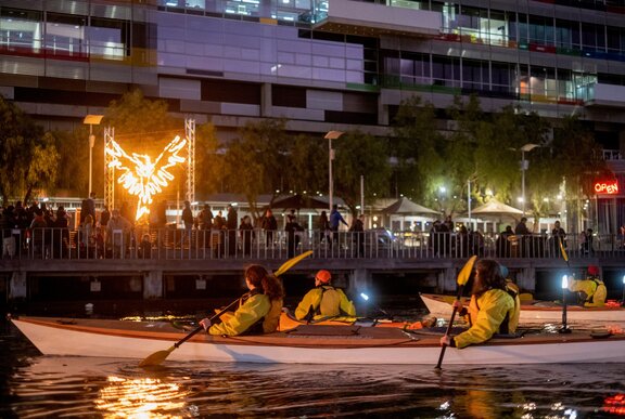 People in kayaks on the water next to a busy walkway with a fire installation in the background.