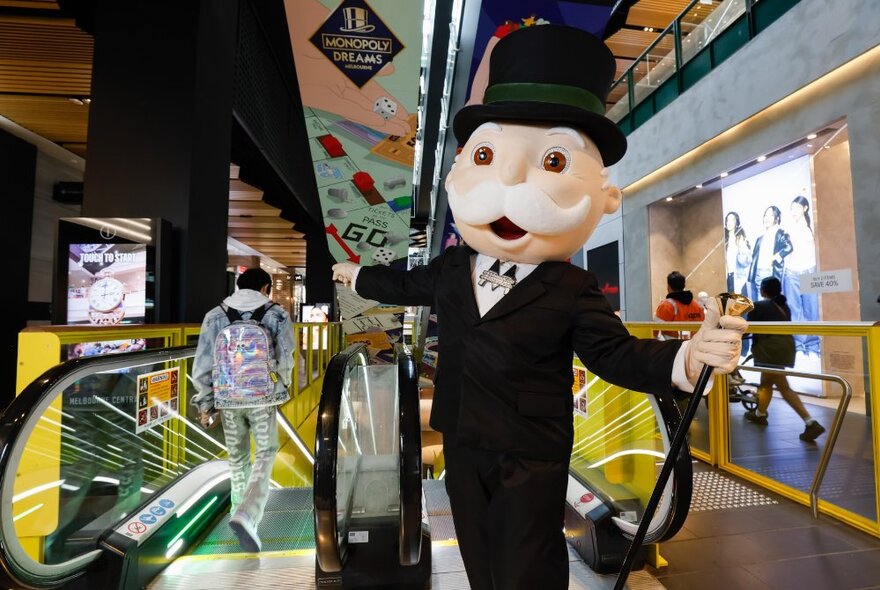 Mr Monopoly character wearing black suit and hat, standing in front of escalators in Melbourne Central.