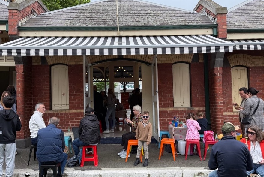 People dining on plastic stools at a cafe under the verandah of a terrace house with striped awning.
