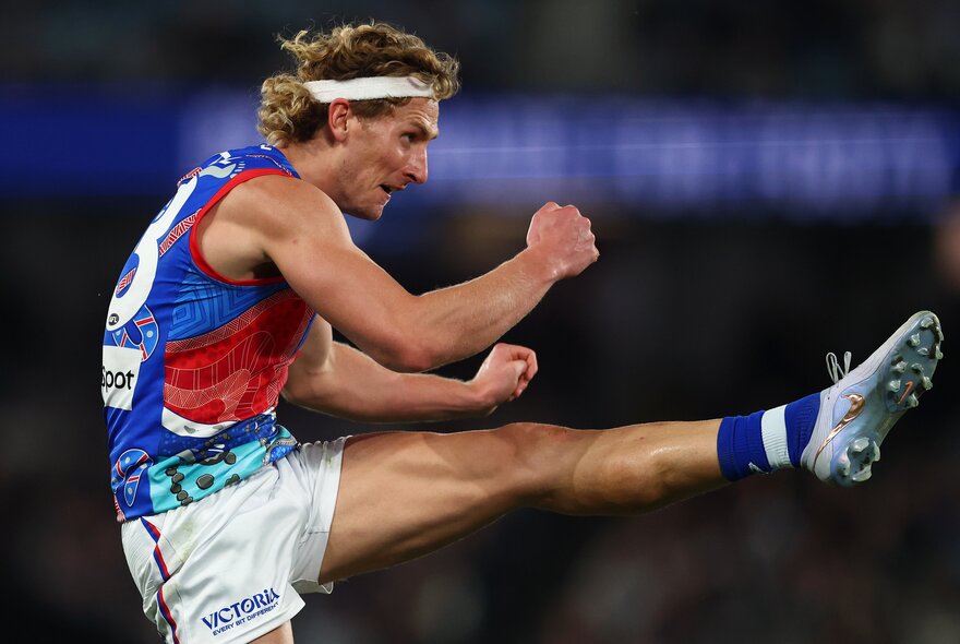 A Western Bulldogs AFL player with his leg raised to hip height having just kicked a football. 