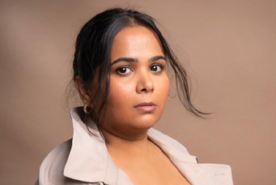 Comedian Sumukhi Suresh looking serious with her hair up but wispy around the face against a camel coloured background. 