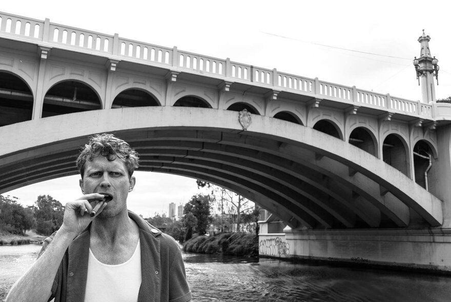 Comedian David Quirk with a cigar in his mouth, standing on the banks of the Yarra River with a bridge in the background.